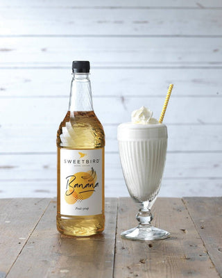 Bottle of Sweetbird Banana syrup next to a glass of banana milkshake on a wooden surface.