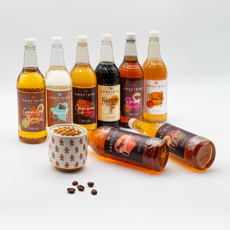 Assorted bottles of Sweetbird syrups on a white background with a small cup and coffee beans.