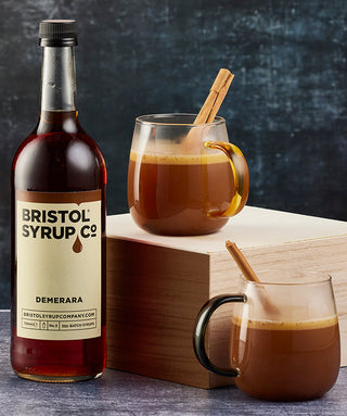 Bottle of Bristol Syrup Co. Demerara syrup with two glass cups of cinnamon-flavoured coffee, garnished with cinnamon sticks, on a wooden block against a dark background