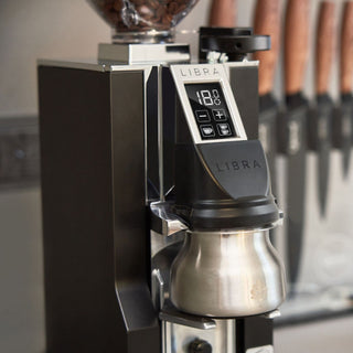 Eureka Mignon Libra 65 grinder with stainless steel dosing cup and digital display, shown in a café setting