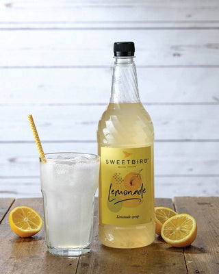 Bottle of Sweetbird lemonade with a glass of lemonade and lemons on a wooden surface.